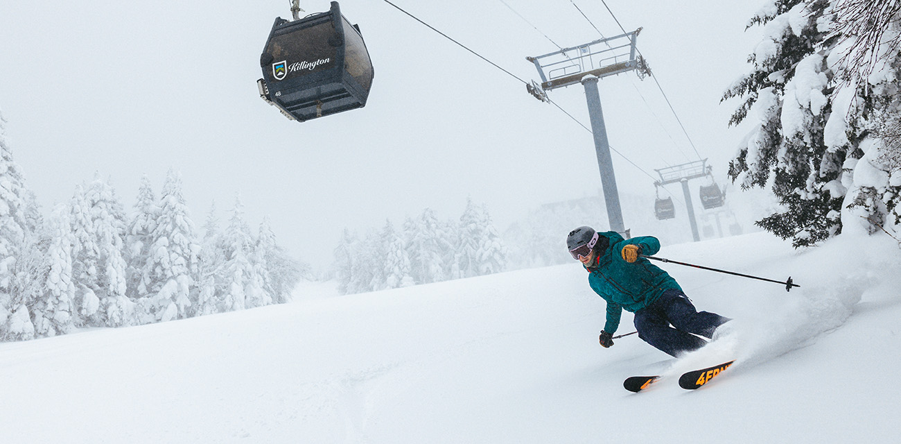 Skier carving through fresh snow beneath a gondola on a snowy mountain.